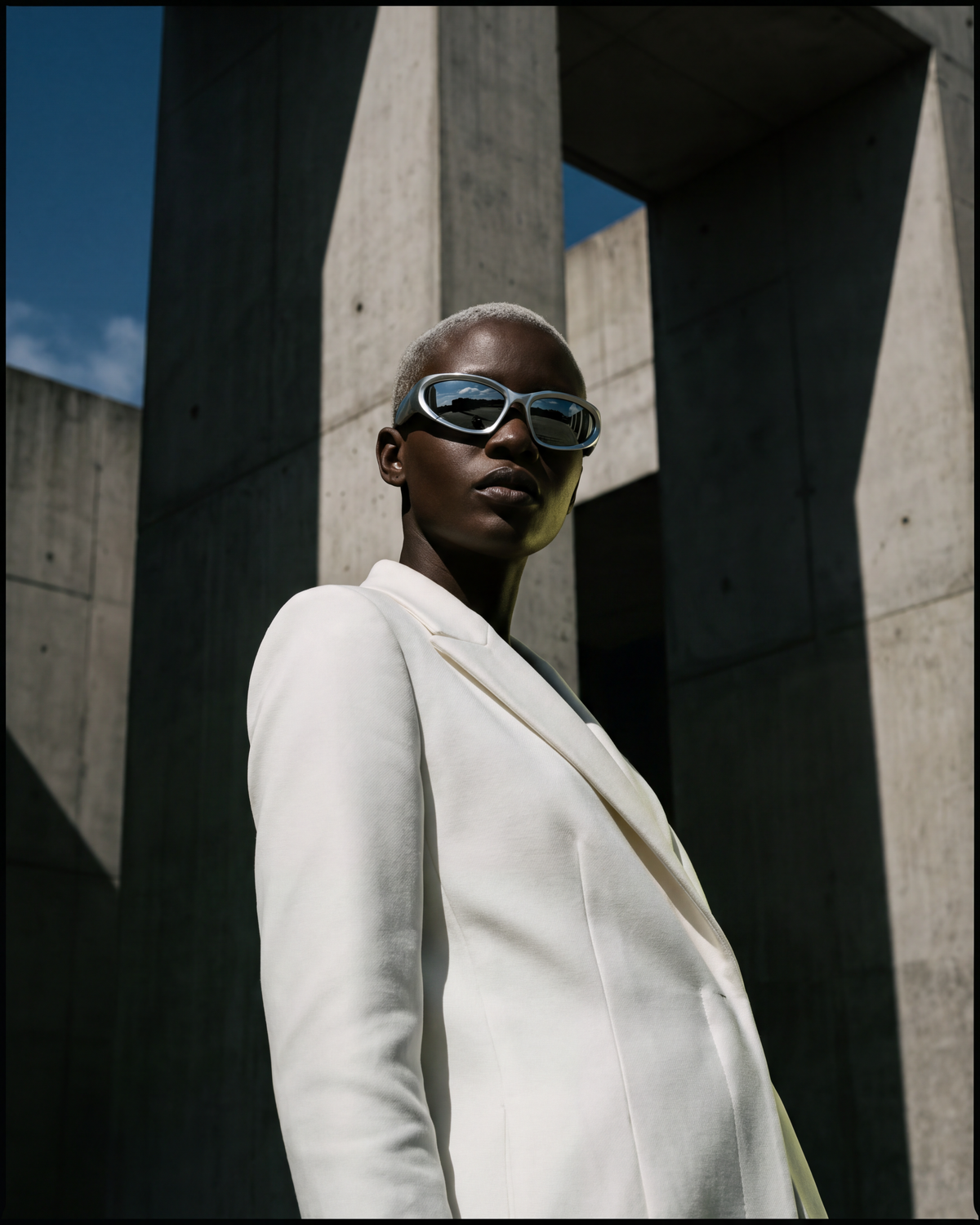 Model in chrome mirror shield sunglasses and white coat against concrete architecture